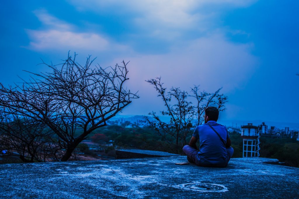 A man sitting on a roof looking out over a city with 2 trees that are bare to the bark and an evening blue haze in the sky.