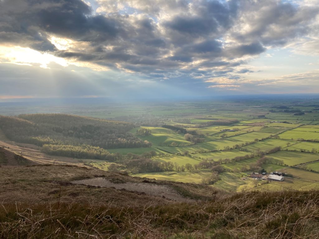 Image of the sun creeping through the clouds over a view of fields and hills.