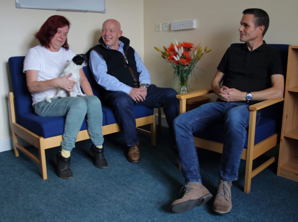 Three people talking in a quiet room, one with a dog.