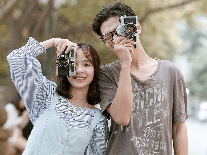 A couple smiling whilst holding cameras up in front of their faces.