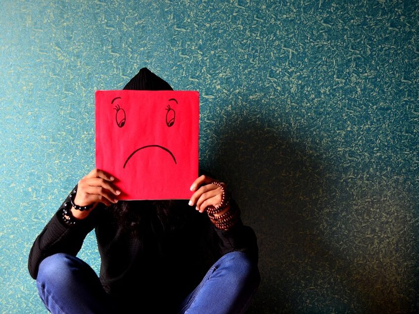 Person sat with a red piece of card that has a sad face on it, covering their face.